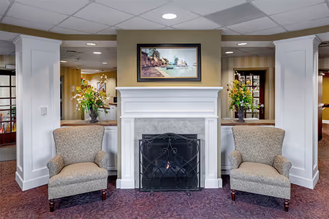 A cozy interior seating area featuring two patterned armchairs placed on either side of a white fireplace with a black metal screen. Above the fireplace hangs a framed painting of a lakeside scene with trees and a small boat. On both sides of the fireplace, there are large vases with colorful floral arrangements. The room has a carpeted floor and a ceiling with recessed lighting.