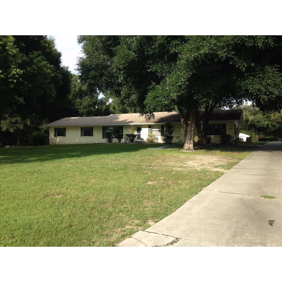 Single-story ranch-style house set back on a large grassy lawn with trees and a driveway.