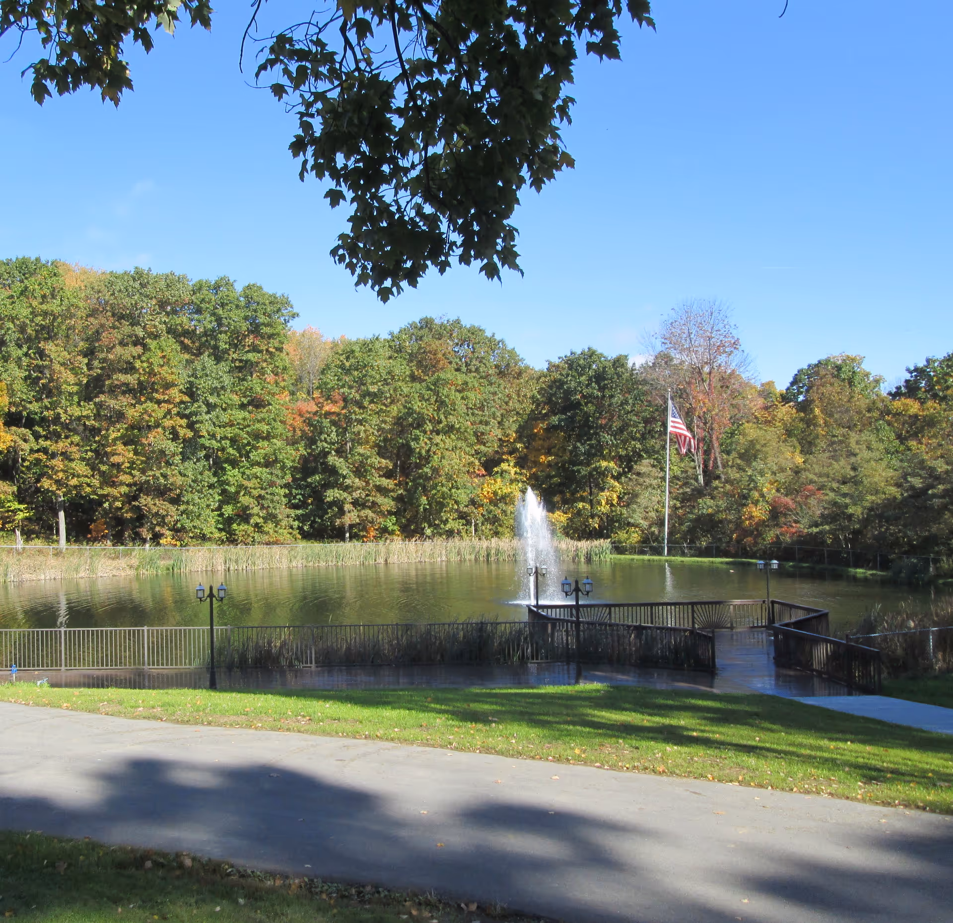 A pond with a central fountain, a dock and an American flag, surrounded by trees under a clear blue sky.
