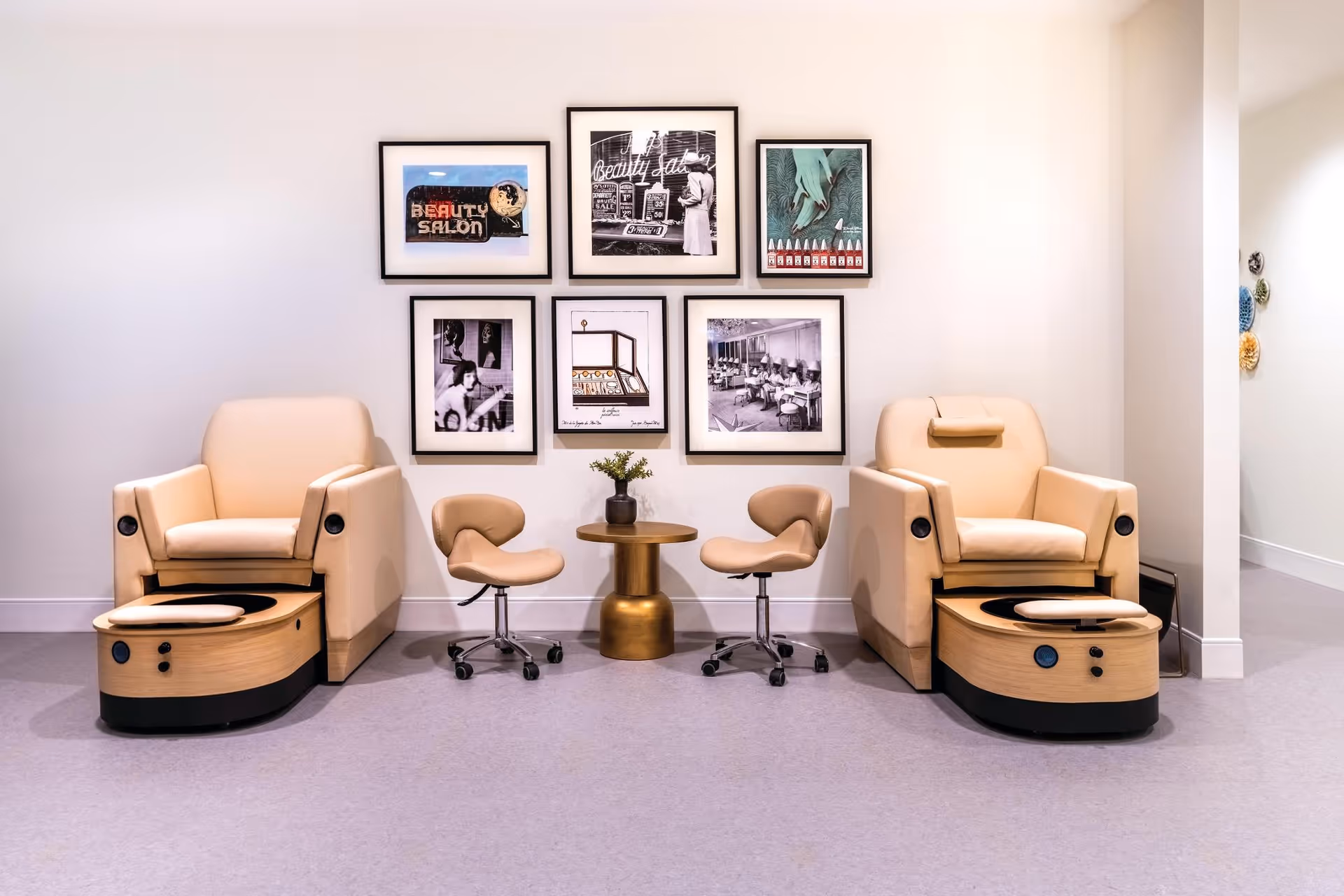 Two beige pedicure chairs with footbaths, two stools and framed artwork on a white wall in a salon-style interior.