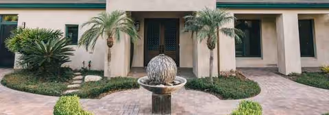 Front entrance of a beige building with palm trees and a central fountain sculpture.