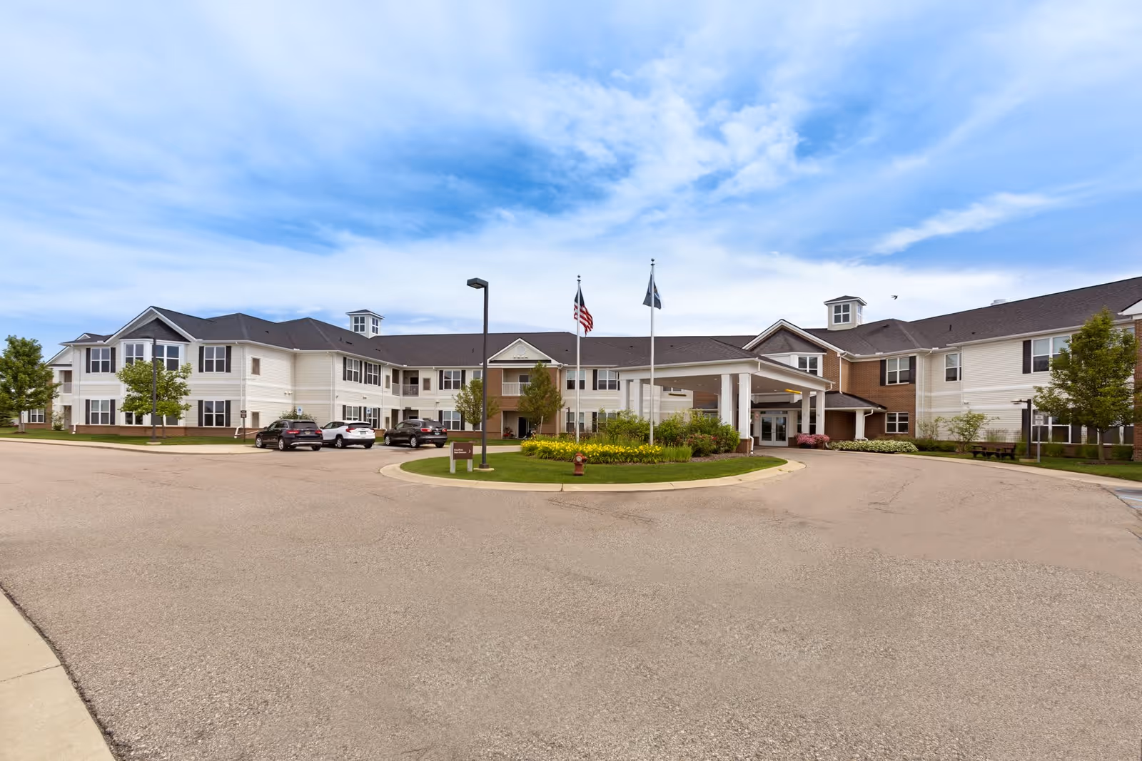 Front exterior view of Olivia's Assisted Living facility showing a large two-story building with a covered entrance, several parked cars, two flagpoles with flags, and a landscaped roundabout with flowers and greenery under a partly cloudy sky.