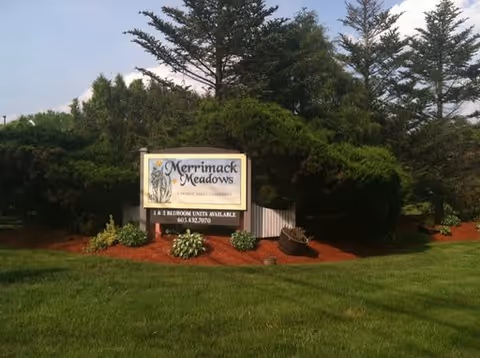 A sign for Merrimack Meadows surrounded by green grass, small plants, and trees in the background under a clear sky.