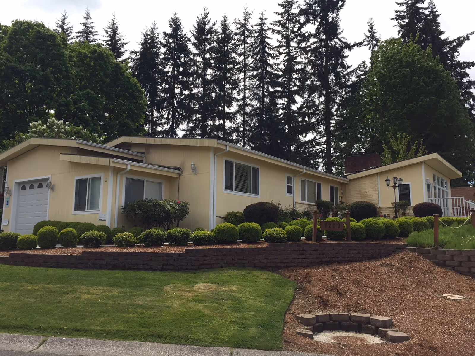 Exterior view of a single-story yellow building with multiple windows, a garage door, and well-maintained landscaping including trimmed bushes and a green lawn. Tall evergreen trees are visible in the background under a cloudy sky.