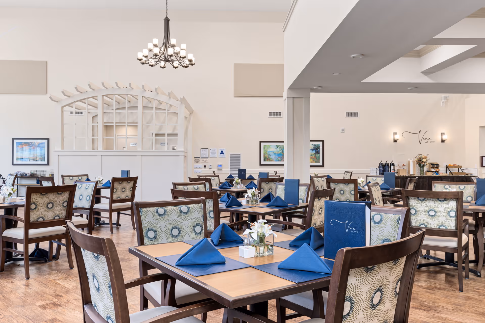 Bright dining room in a senior living facility with multiple wood tables set with blue napkins and floral centerpieces.