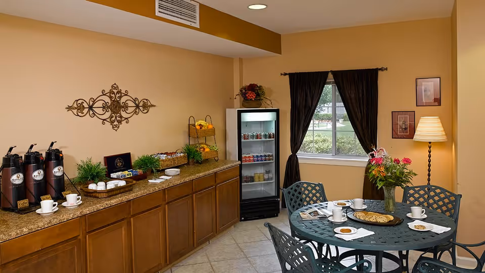 A cozy dining area with a round metal table set with cups, plates of cookies, and a vase of flowers. Behind the table is a window with dark curtains, a floor lamp, and framed pictures on the wall. To the left, there is a countertop with coffee dispensers, cups, plates, baskets of fruit, and a small refrigerator stocked with beverages.