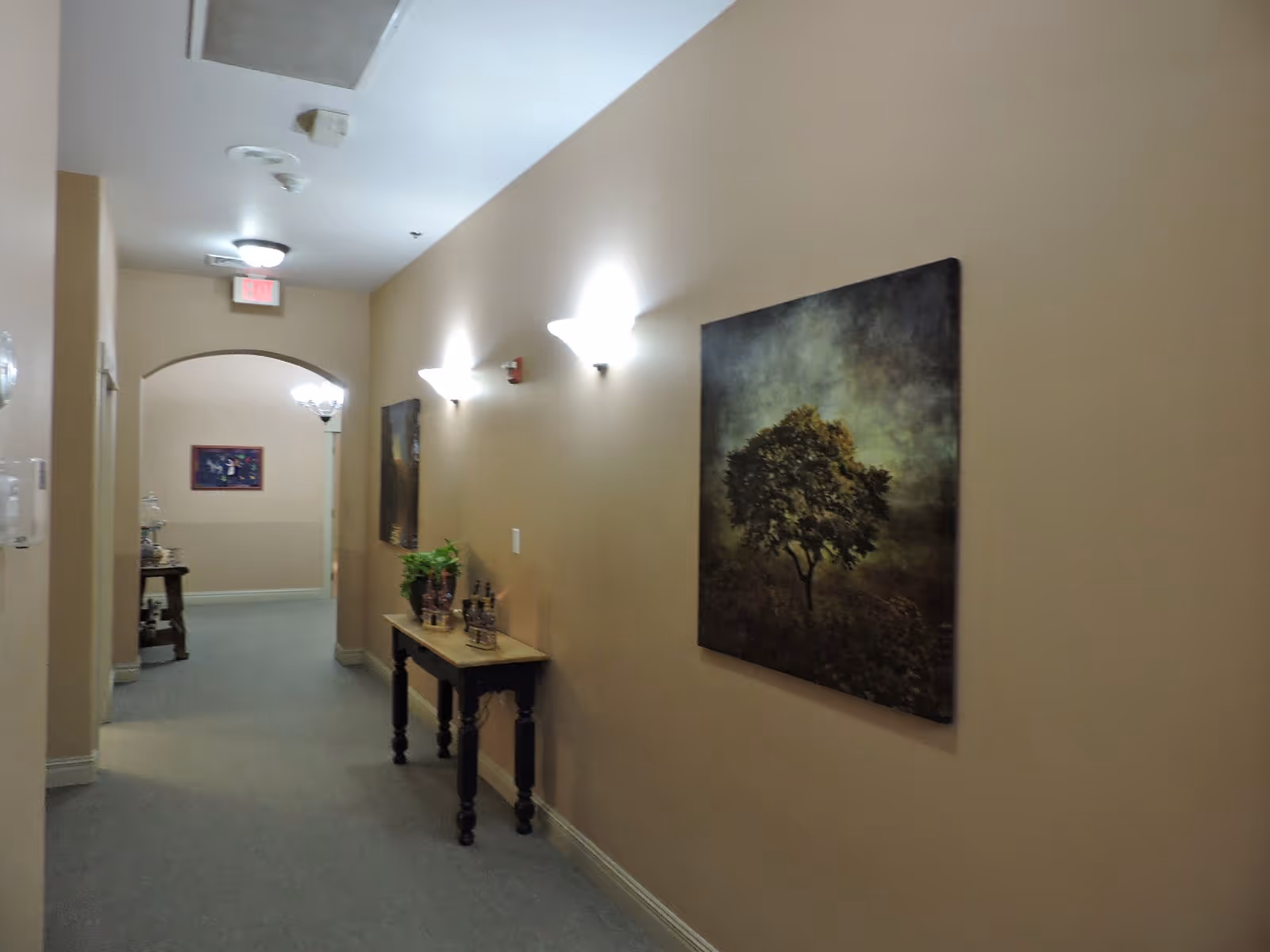 A softly lit hallway with beige walls and carpeted floor. On the right wall, there is a large painting of a tree. Below the painting is a narrow wooden table with decorative items and a small plant. The hallway leads to an arched doorway with another piece of artwork visible in the background.