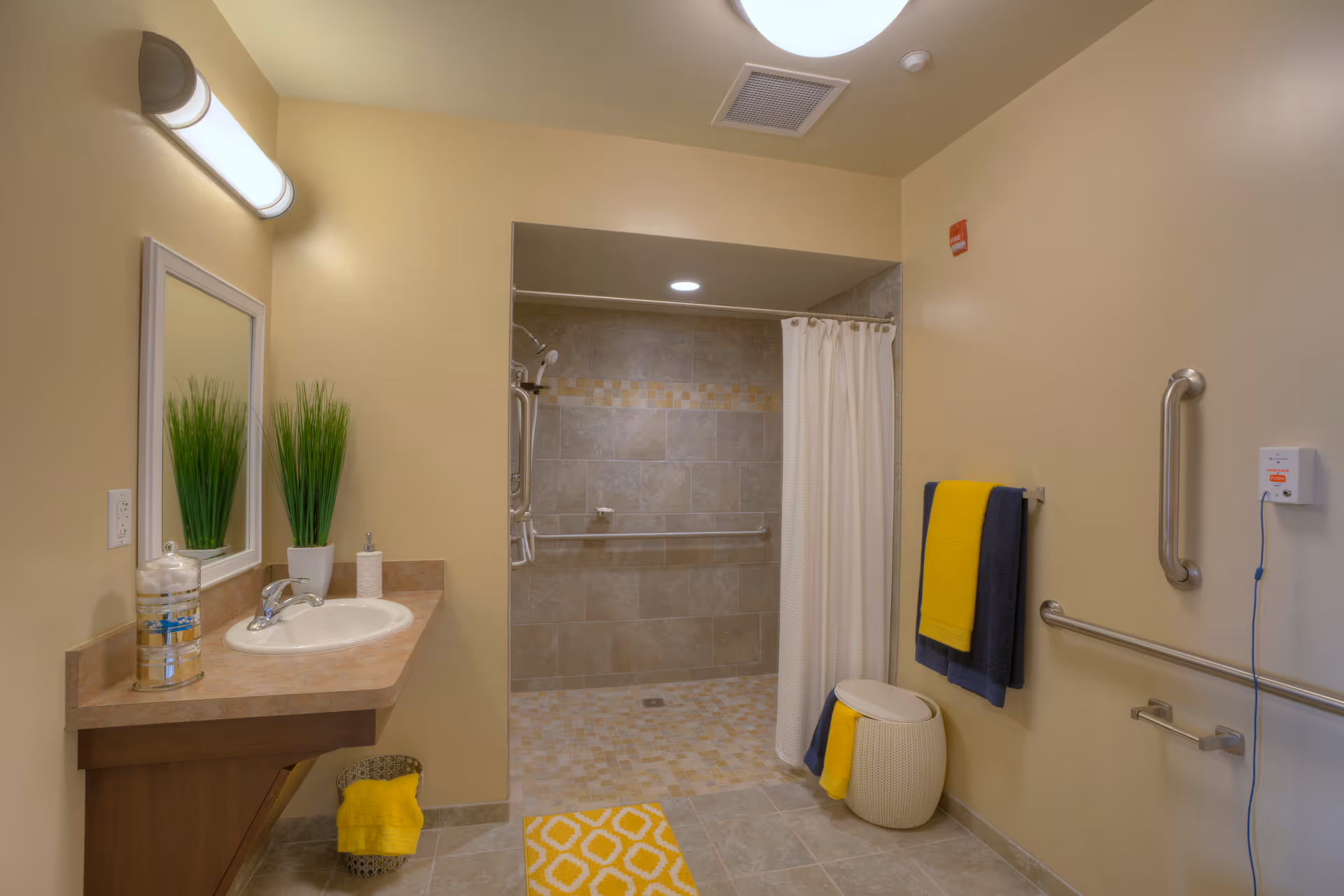 Accessible bathroom with a walk-in tiled shower, sink vanity, grab bars, and yellow towels and rug.