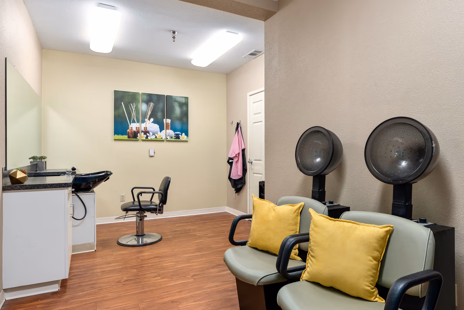Interior view of a salon area in Spring Creek Assisted Living featuring two hair drying chairs with yellow pillows, a black salon chair in front of a wash basin, a countertop with a small plant and decorative item, and a three-panel wall art depicting spa items on a beige wall.