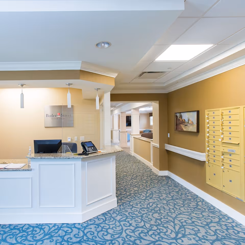 Reception area of Butler Street Senior Living facility with a white front desk, computer, phone, and a clear protective screen. The floor is covered with a blue patterned carpet, walls are painted beige, and there are yellow mailboxes mounted on the right wall. A hallway extends beyond the reception area with seating visible in the distance.