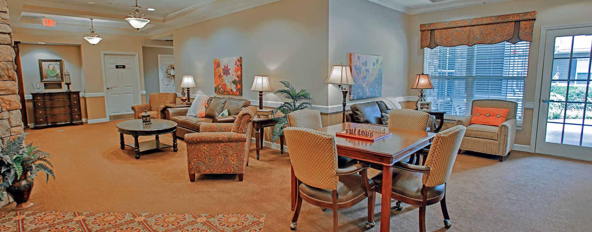 A cozy living room area in a senior living facility with multiple seating options including sofas and armchairs. There is a wooden table with four chairs and a chessboard on top. The room is well-lit with several table lamps and natural light coming through large windows with blinds. The walls are decorated with framed artwork, and there are plants placed around the room. A door with glass panels leads outside to a garden or patio area.