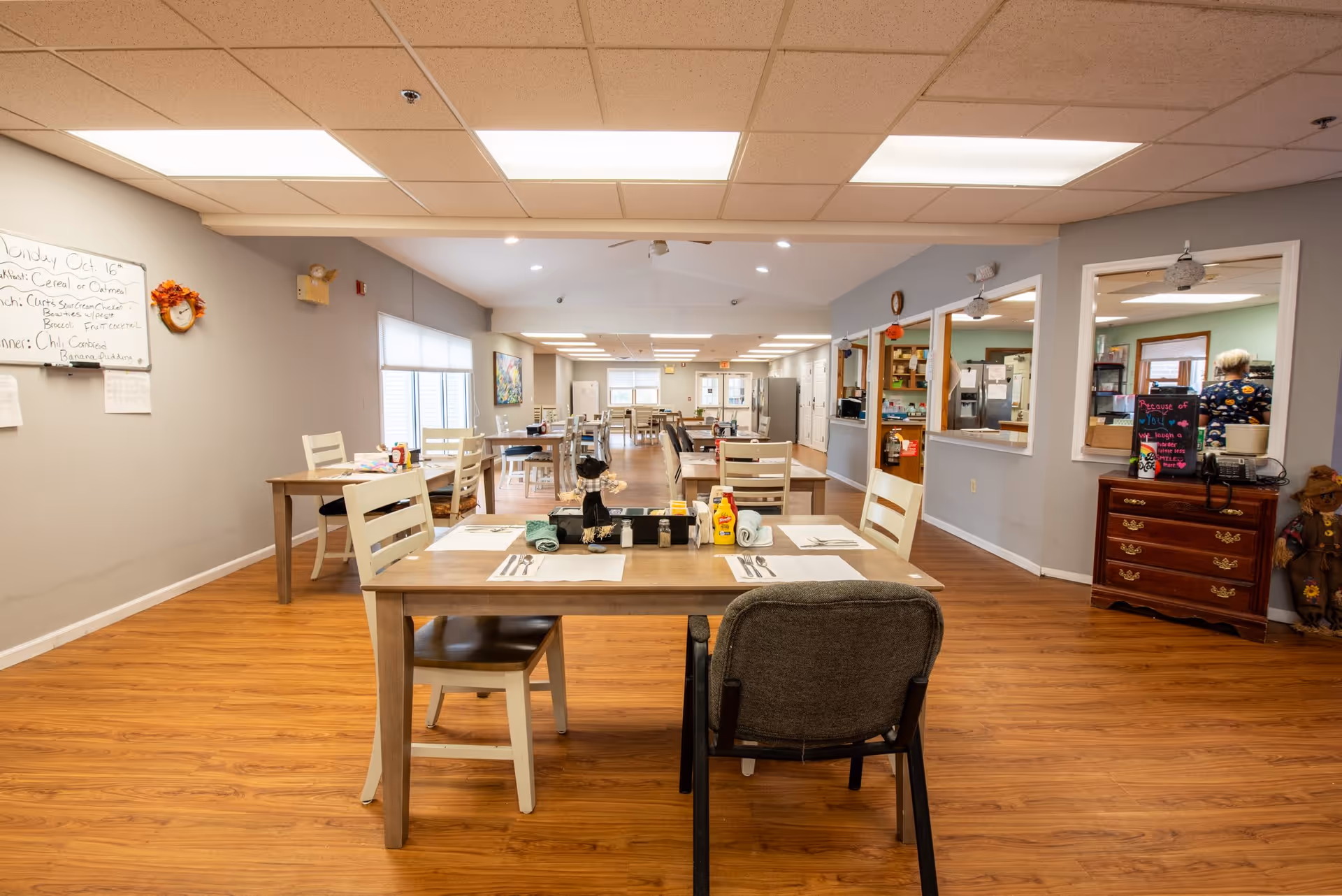 A spacious dining room with multiple wooden tables and chairs arranged neatly. Each table is set with placemats, utensils, condiments, and napkins. The room has wood flooring, gray walls, and a white ceiling with fluorescent lighting. A whiteboard on the left wall displays the day's menu. There is a kitchen area visible through windows on the right side, and a person is seen inside the kitchen. The room has large windows allowing natural light to enter.