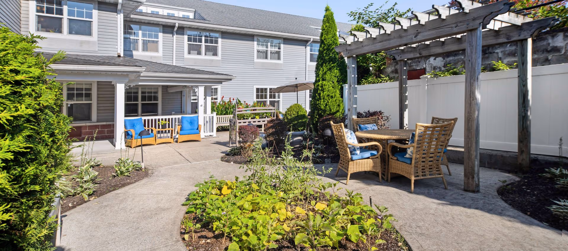 Sunlit courtyard with patio seating, wicker chairs and a wooden pergola in front of a two-story building.