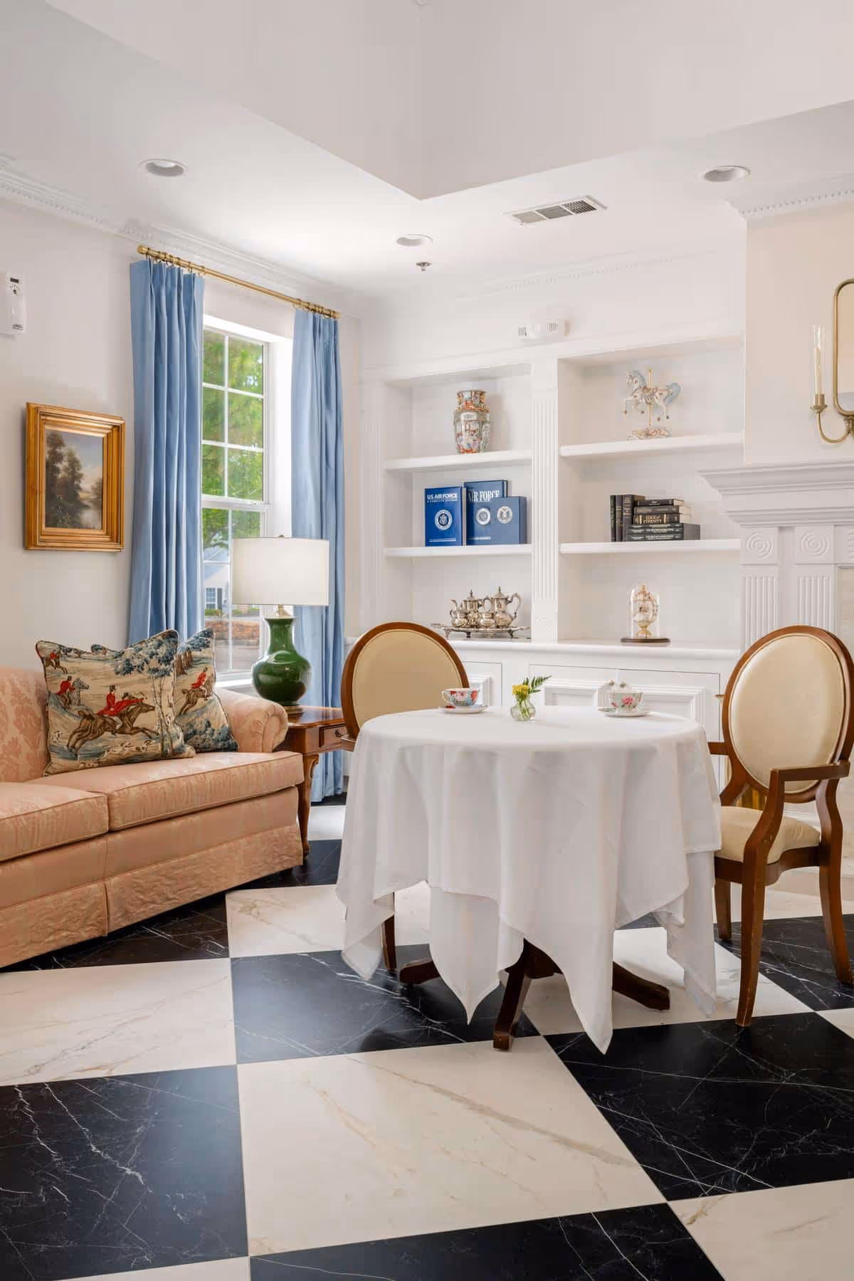 A cozy sitting area in Cumberland Hills Assisted Living featuring a round table covered with a white tablecloth, two upholstered chairs, a peach-colored sofa with decorative pillows, a green table lamp, blue curtains framing a window, and built-in white shelves with decorative items and books.