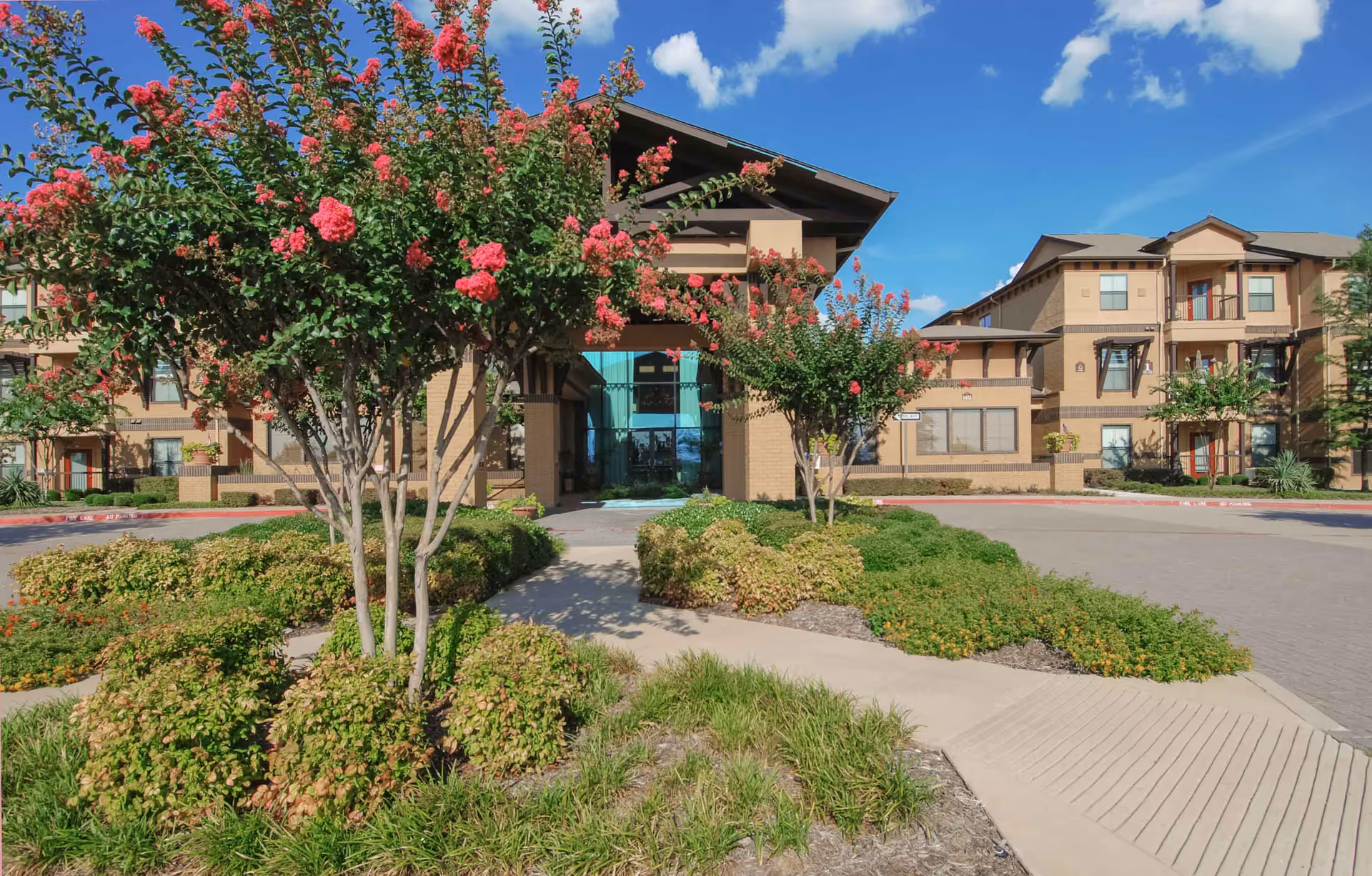 Exterior view of Parkview in Allen senior living facility showing a landscaped entrance with flowering trees and bushes, a paved driveway, and a multi-story building with balconies under a blue sky with scattered clouds.