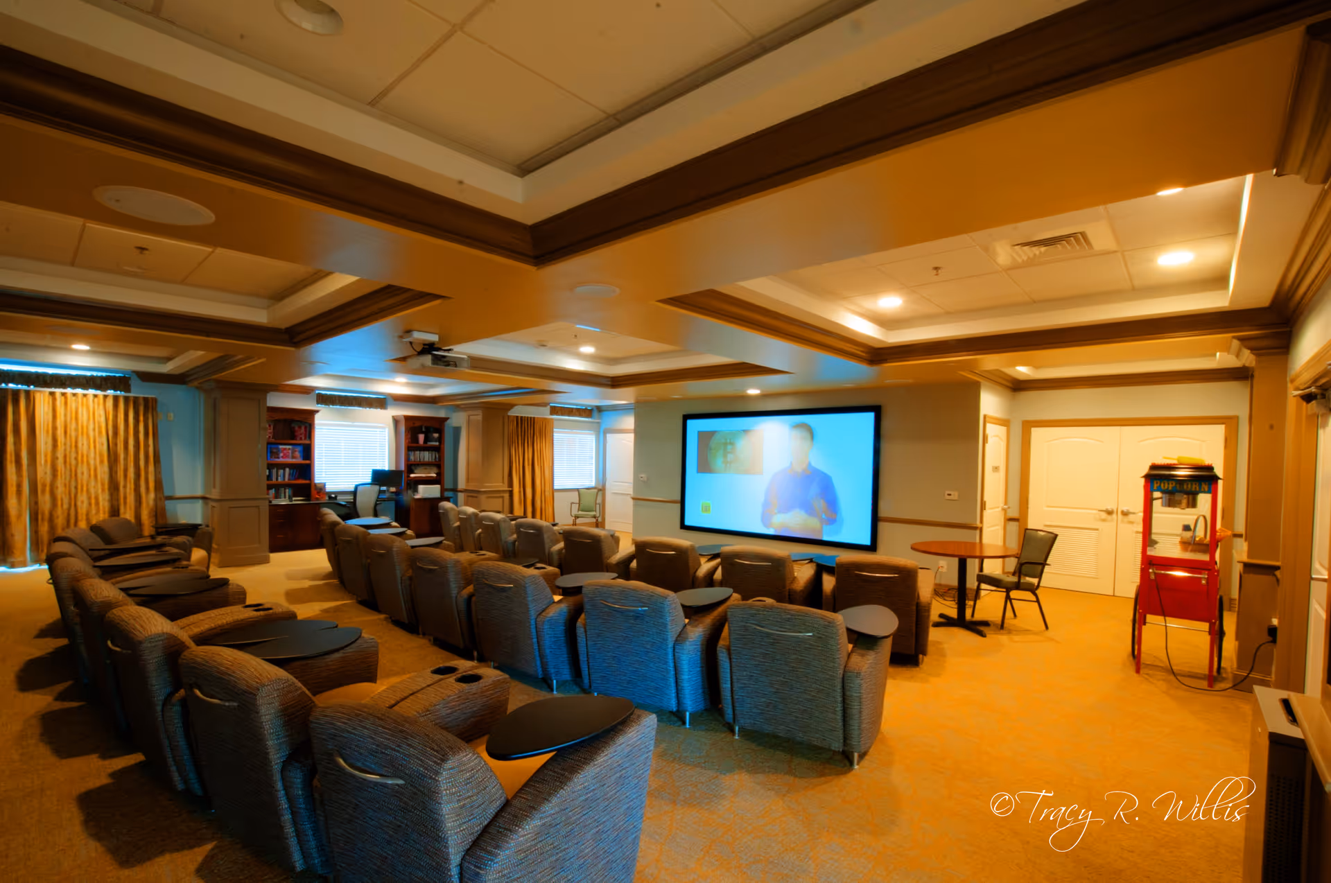 A cozy media room with multiple rows of cushioned recliner chairs facing a large screen on the wall. The room has warm lighting, wood trim on the ceiling, and windows with curtains. There is a popcorn machine and a small table with chairs near the back of the room.