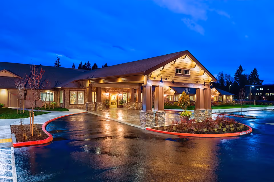 Exterior view of The Hampton at Salmon Creek Memory Care Community building at dusk, featuring a covered entrance with stone pillars, warm lighting, wet pavement, landscaped areas with small trees and shrubs, and a clear blue sky.