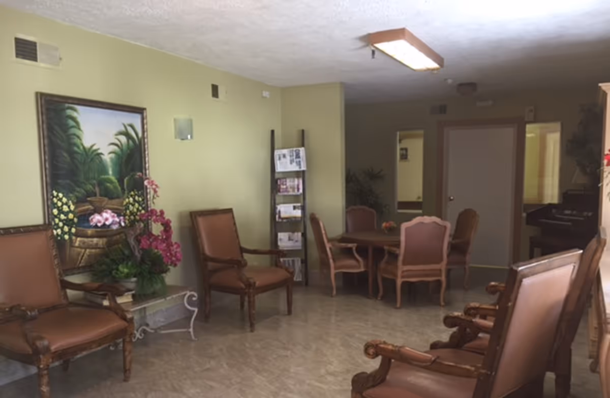 A common area with several brown cushioned wooden chairs arranged around a small round table. There is a painting of a garden scene on the light green wall, a small table with a flower arrangement, and a magazine rack. The room has a tiled floor and a ceiling light fixture.