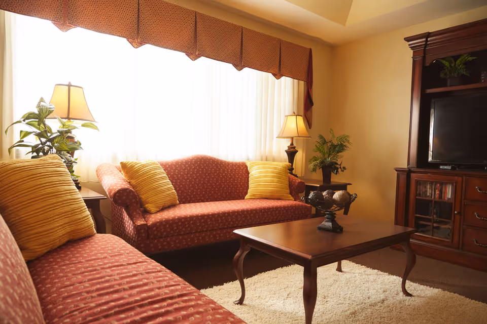 A cozy living room with two red patterned sofas adorned with yellow cushions, a wooden coffee table with decorative items, two table lamps on side tables, potted plants, and a wooden entertainment center with a TV and shelves.