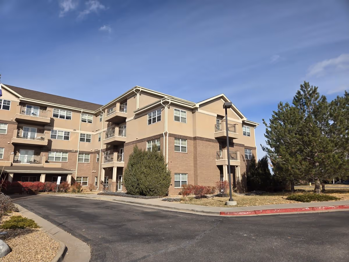 Exterior front view of a multi-story senior living building with balconies, a driveway, and landscaping under a blue sky.