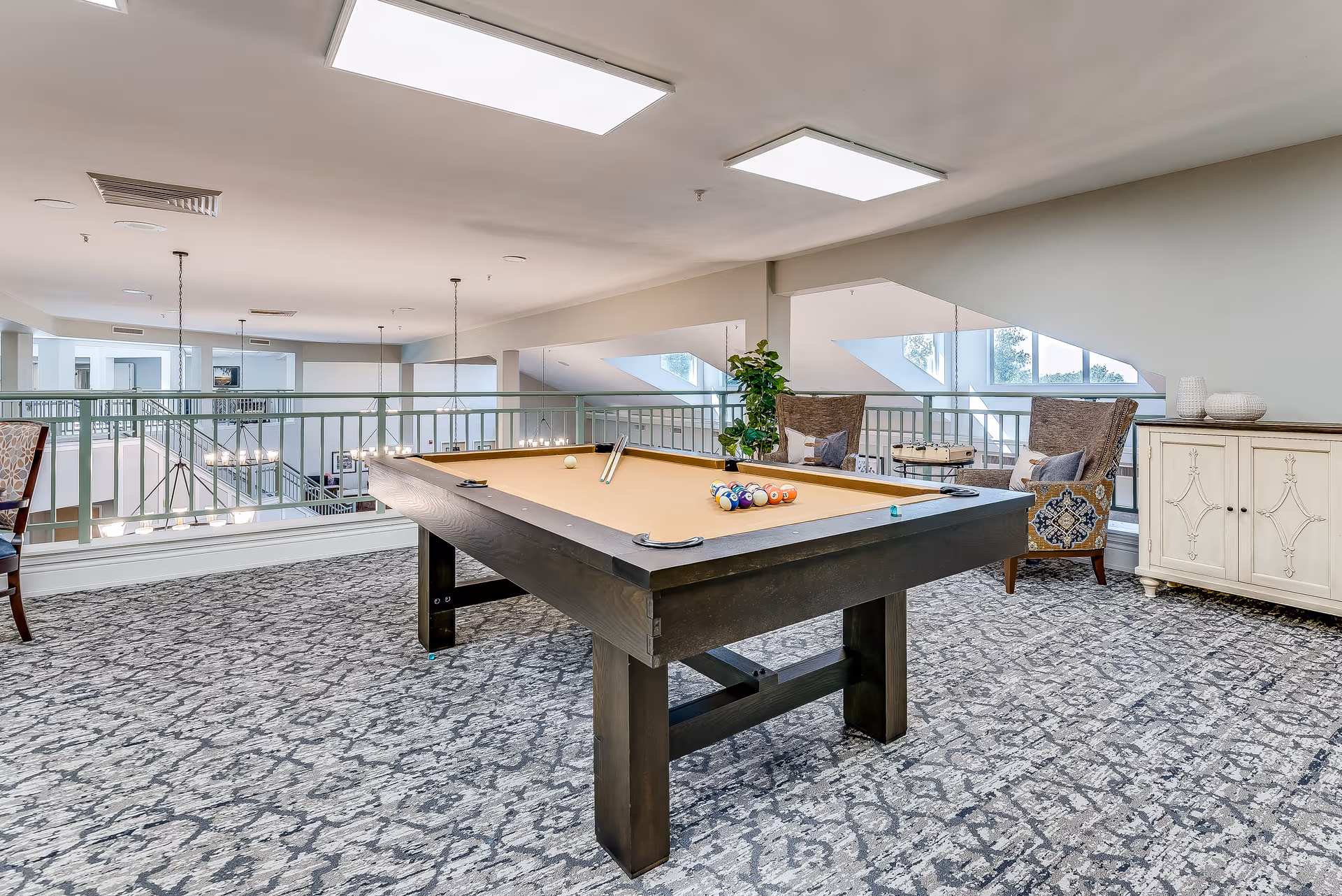 Well-lit common lounge area with a pool table, chairs, and a decorative cabinet overlooking an atrium.