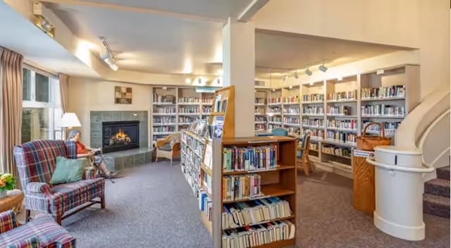 A cozy library room in a senior living facility with bookshelves filled with books, a fireplace with a fire burning, comfortable plaid armchairs, a person sitting and reading near the fireplace, and a spiral staircase leading upstairs.