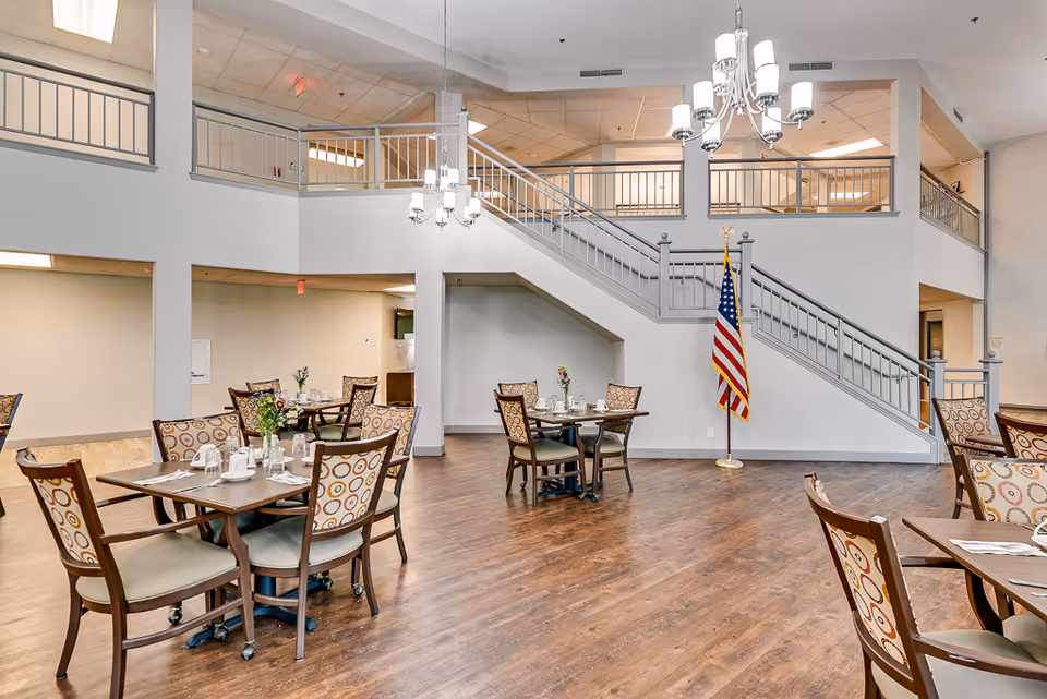 A spacious dining area in a senior living facility with several tables and chairs arranged neatly. Each table is set with white cups, plates, and silverware, and decorated with small flower arrangements. The room features wooden flooring, a staircase with metal railings leading to an upper level, and an American flag positioned near the staircase. Two chandeliers hang from the ceiling providing lighting.