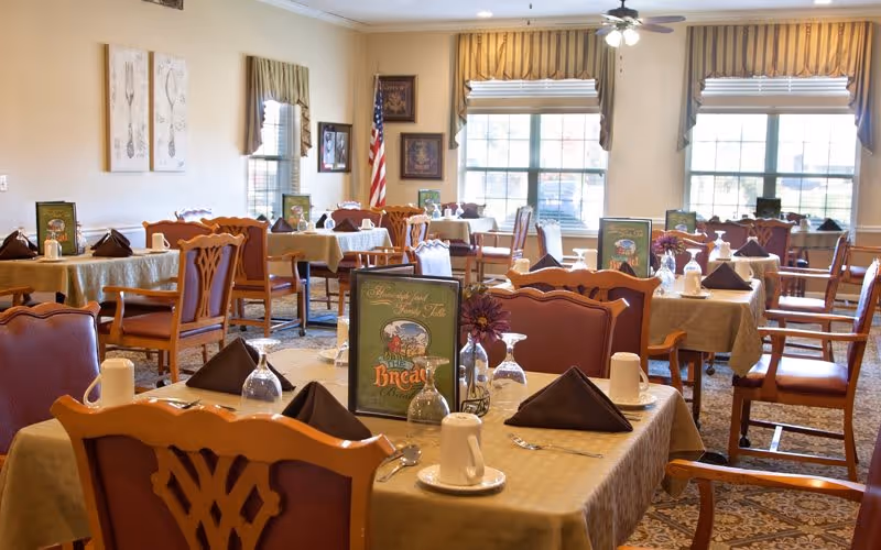 A bright dining room in a senior living community with multiple tables covered in beige tablecloths, each set with brown napkins, white cups, upside-down wine glasses, and menus. The room has large windows with striped valances, framed pictures on the walls, and an American flag in the corner. Wooden chairs with cushioned seats surround the tables.