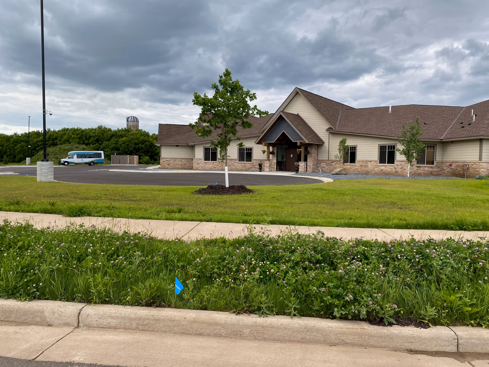 Exterior view of a single-story senior living facility building with beige siding and stone accents, a peaked roof, a small tree in front, a circular driveway, and a parked van in the background under a cloudy sky.