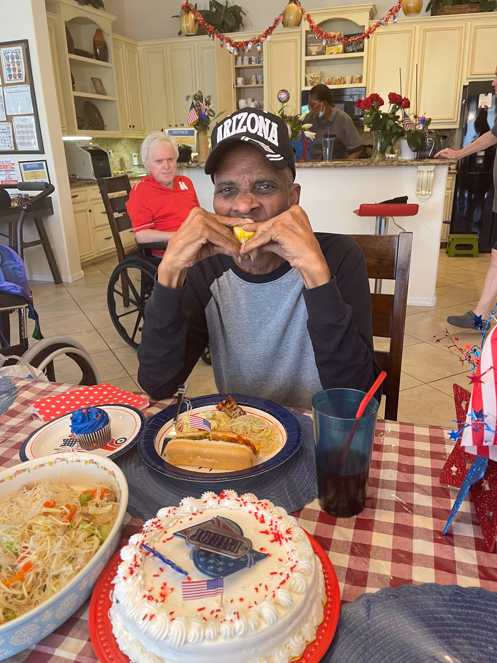 An elderly man wearing an Arizona cap is sitting at a table with a checkered tablecloth, eating corn on the cob. On the table in front of him are a hot dog, a cupcake with blue frosting, a bowl of noodles with vegetables, a decorated white cake with red sprinkles and a small American flag, and a dark drink with a red straw. In the background, there is a kitchen area with cabinets, a counter decorated with red garland, and two other elderly individuals, one in a wheelchair and another standing near the counter.