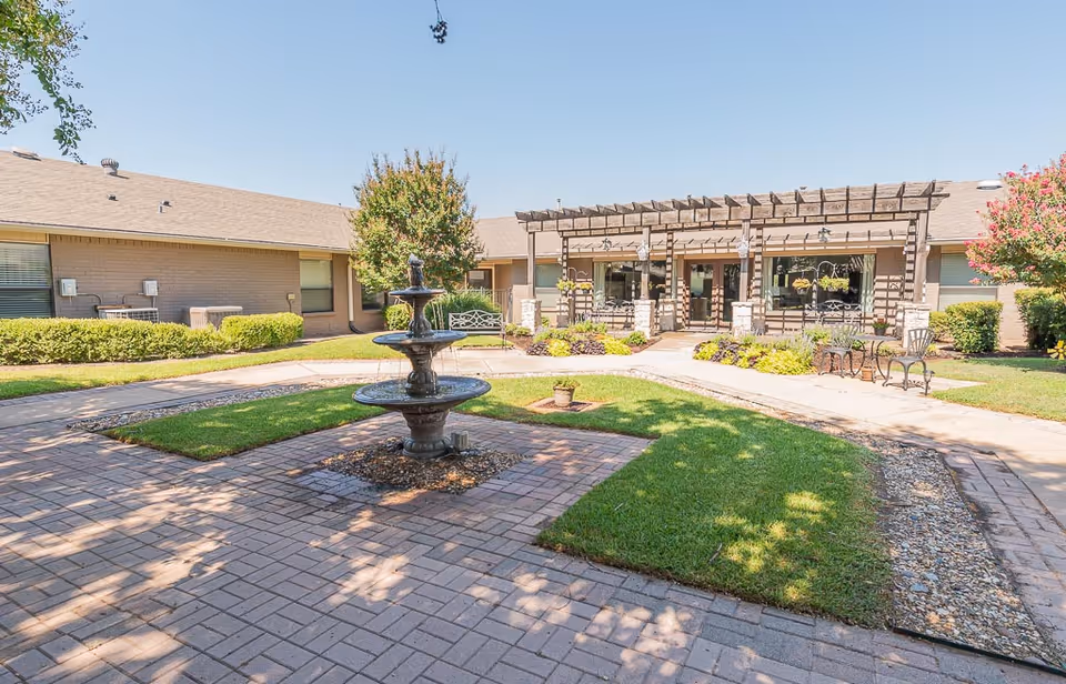 Outdoor courtyard area of a senior living facility featuring a three-tiered water fountain in the center, surrounded by paved walkways, green grass, shrubs, and trees. The building has a covered patio with hanging plants and outdoor seating under a wooden pergola.
