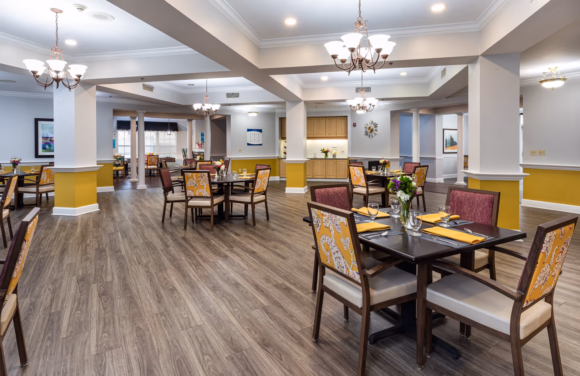 A spacious dining room with multiple tables and chairs arranged neatly. Each table is set with yellow napkins, glassware, and a small floral centerpiece. The room features wood flooring, white walls with a yellow accent, and several chandeliers hanging from the ceiling providing warm lighting. There are decorative columns and framed artwork on the walls.