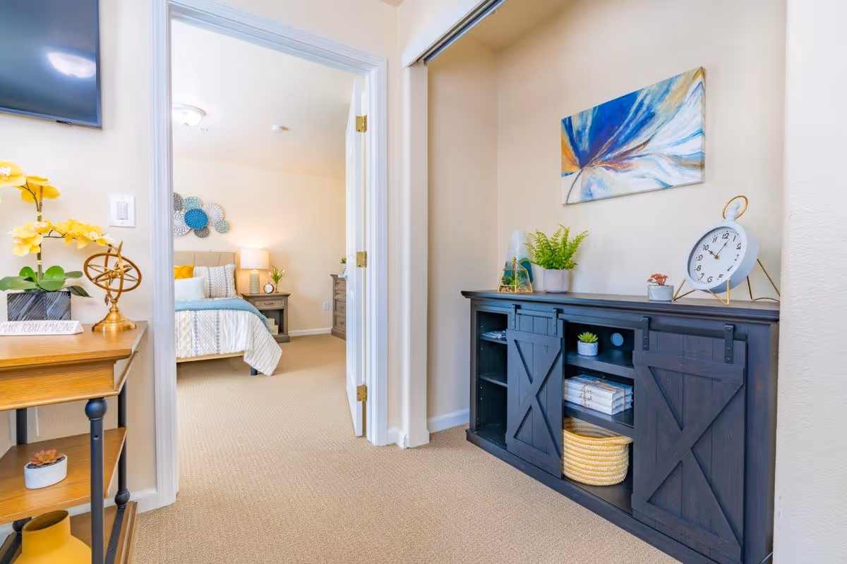 Interior view of a hallway with a dark console table and decorative items, looking into a furnished bedroom through an open doorway.