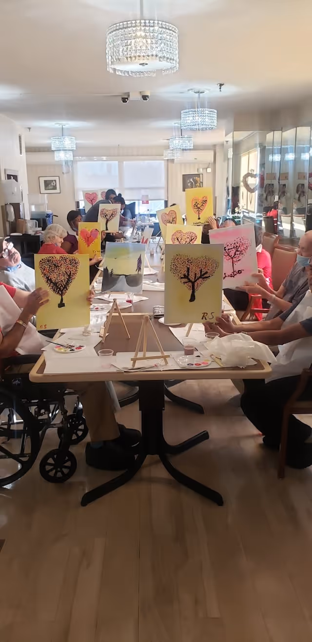 A group of elderly people seated around tables in a well-lit room, each holding up paintings of trees with heart-shaped leaves. The room has modern chandeliers and large windows letting in natural light. Some participants are wearing masks.