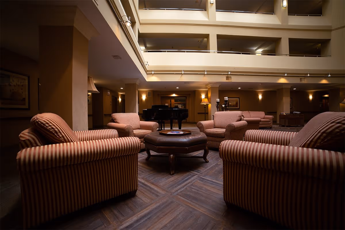 A cozy indoor common area with four striped armchairs arranged around a hexagonal ottoman. In the background, there is a grand piano and additional seating, with warm lighting and beige walls. The space is open to upper floors with railings visible above.