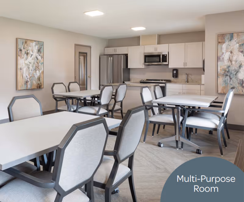A bright multipurpose dining room with tables, chairs, and a kitchenette in the background.