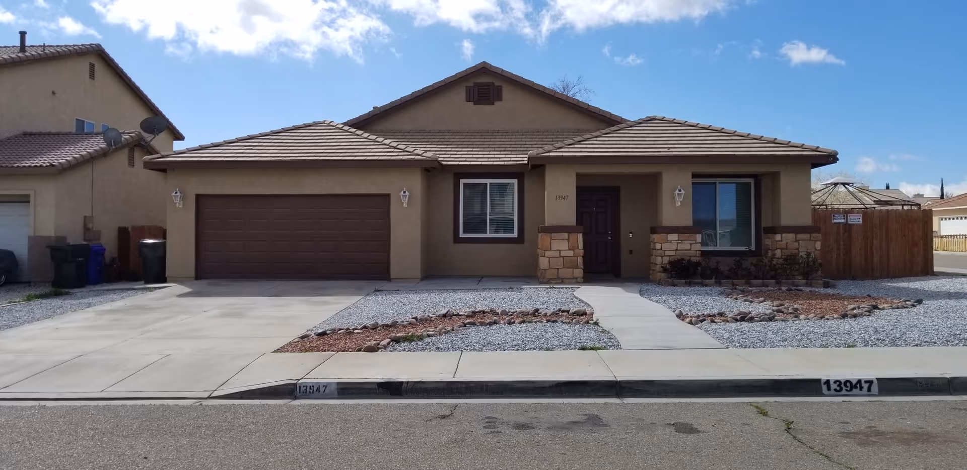 Single-story stucco house with an attached two-car garage, paved driveway, and rock landscaping under a partly cloudy sky.