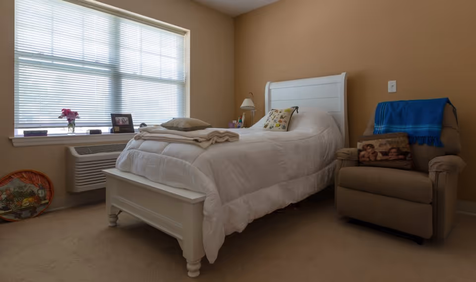 Bedroom with a white bed, beige walls, a large window with blinds, and a recliner chair.