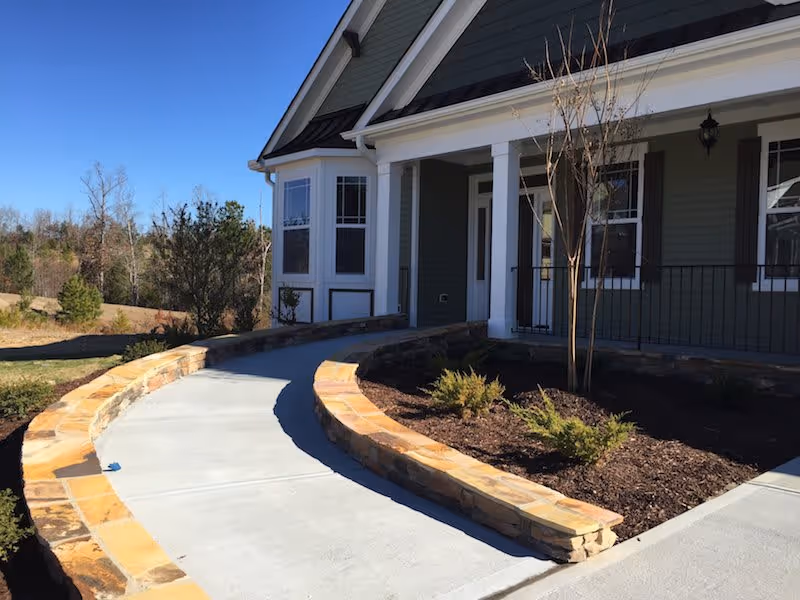 Curved concrete walkway bordered by low stone walls leading to the entrance of a green house with white trim, a small landscaped area with young shrubs and a tree, and a clear blue sky in the background.