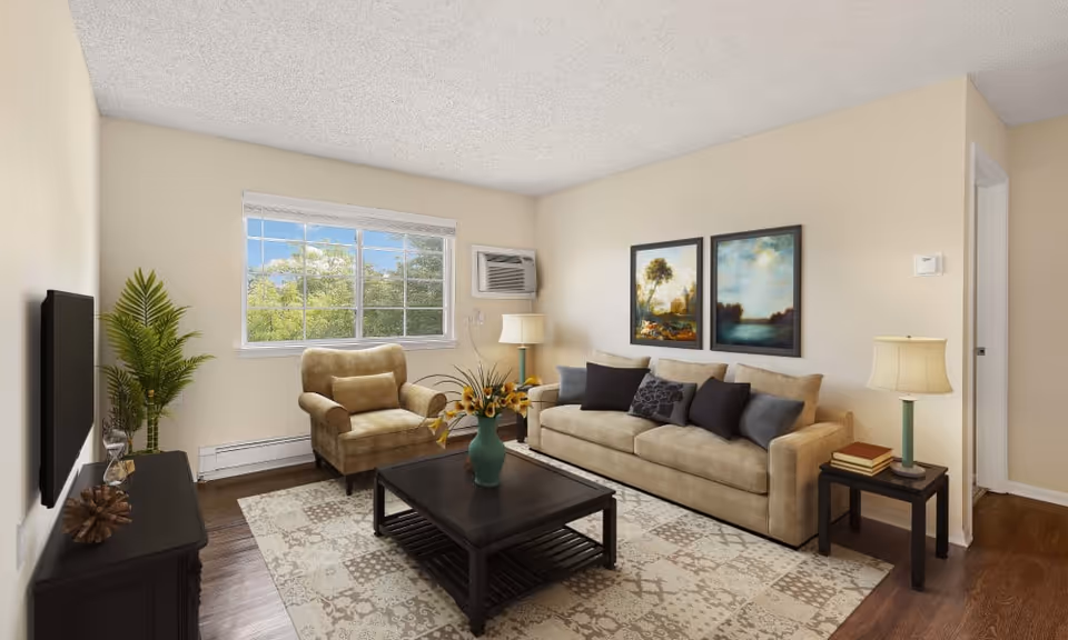A cozy living room with a beige sofa adorned with multiple cushions, a matching armchair, a dark wooden coffee table with a vase of flowers, and a side table with a lamp and books. There is a large window showing green trees outside, two framed landscape paintings on the wall, a flat-screen TV mounted on the opposite wall, and a potted plant near the TV stand.