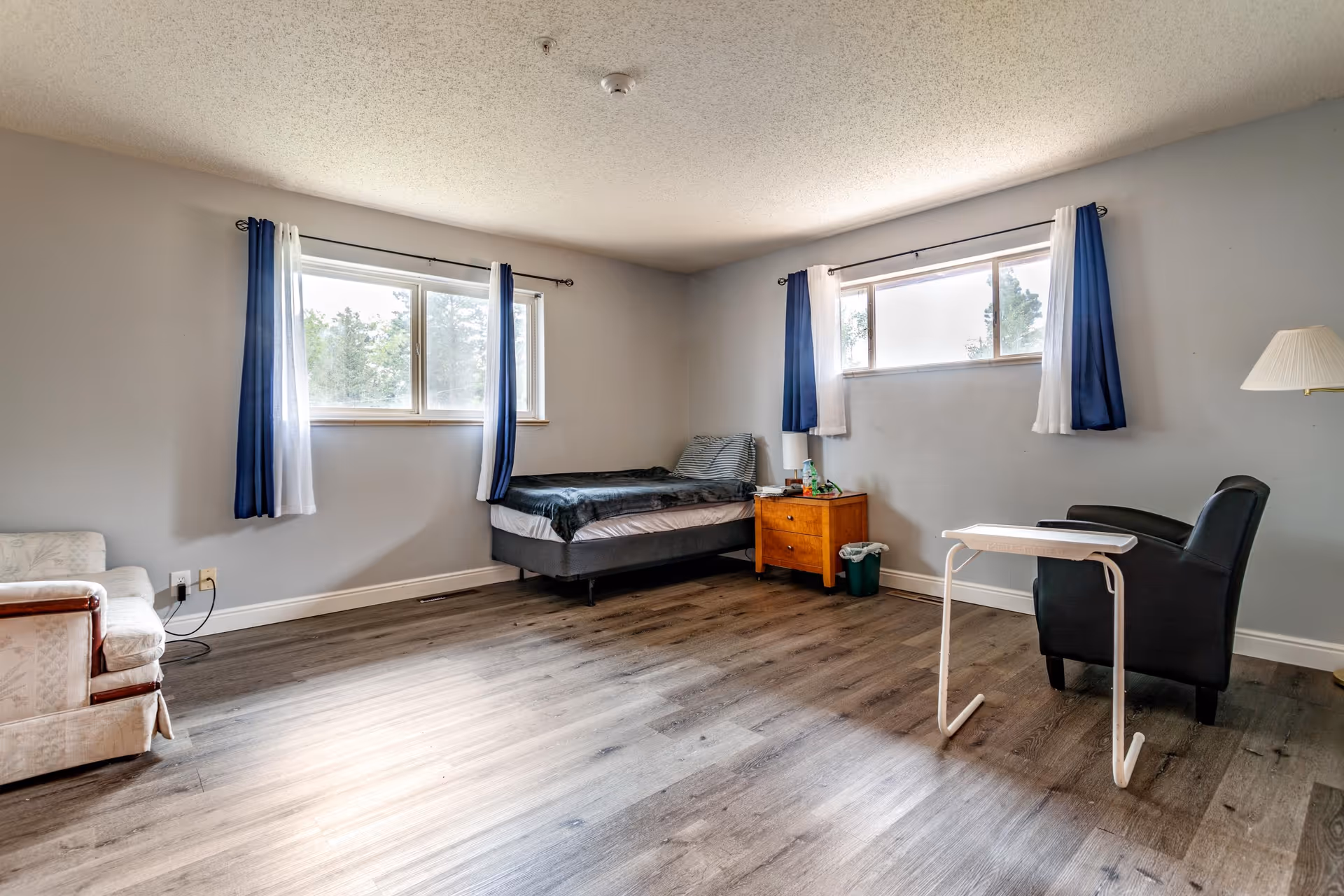 A simple, spacious bedroom with two windows covered by blue and white curtains. The room contains a single bed with a dark blanket, a wooden nightstand with items on it, a black armchair with a small white table in front of it, a floor lamp, and a light-colored upholstered chair. The floor is wood laminate and the walls are painted light gray.