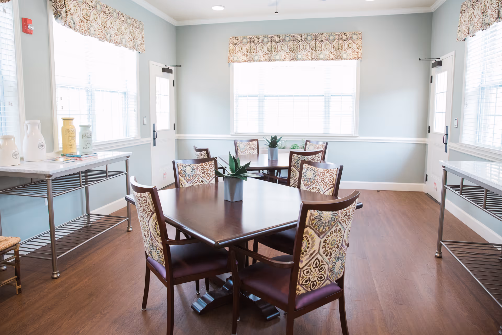 A bright dining room with two dark wooden tables, each surrounded by six chairs with patterned upholstery. The room has light blue walls, large windows with patterned valances, and wooden flooring. There are two metal shelving units along the walls with decorative jars and books on one of them.