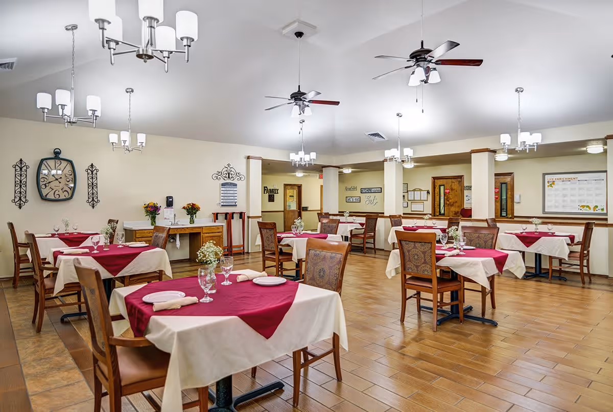 A dining room in a senior living facility with multiple tables covered in white tablecloths and red table runners. Each table is set with plates, glasses, and napkins. The room has wooden chairs, chandeliers, ceiling fans, and wall decorations including a large clock and framed signs. The floor is tiled and the walls are painted light beige.
