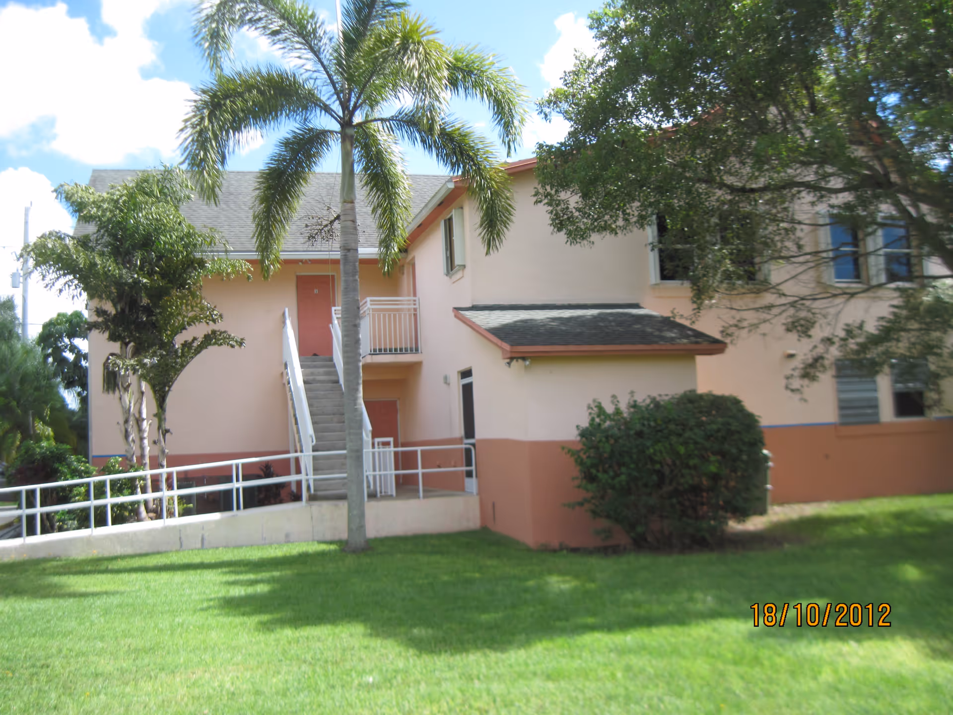 Exterior view of a two-story building with peach and pink walls, a staircase leading to the second floor, surrounded by green grass, palm trees, and other vegetation under a partly cloudy sky.
