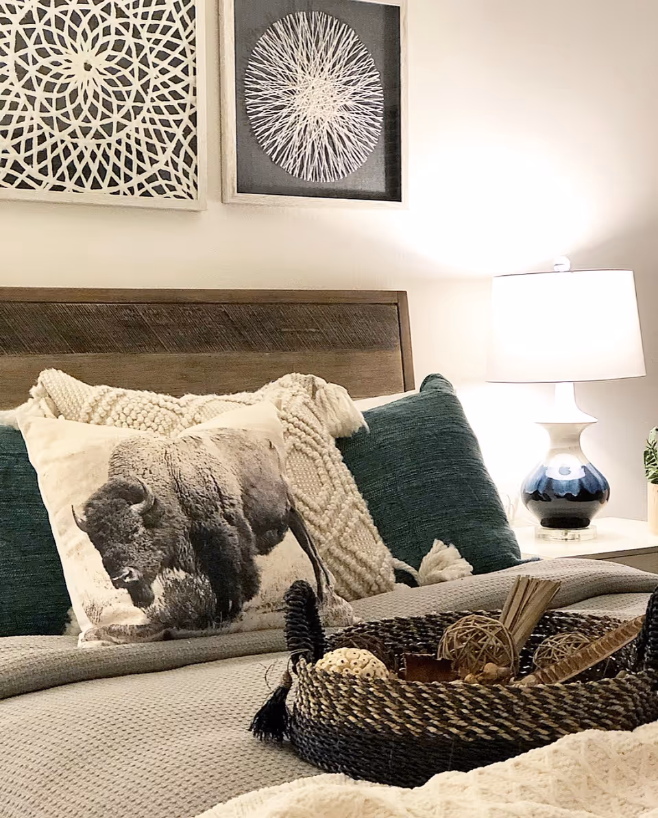 Cozy bedroom scene with a wooden headboard, decorative pillows including a bison-print cushion, a woven tray on the bed, a bedside lamp, and framed wall art above the bed.