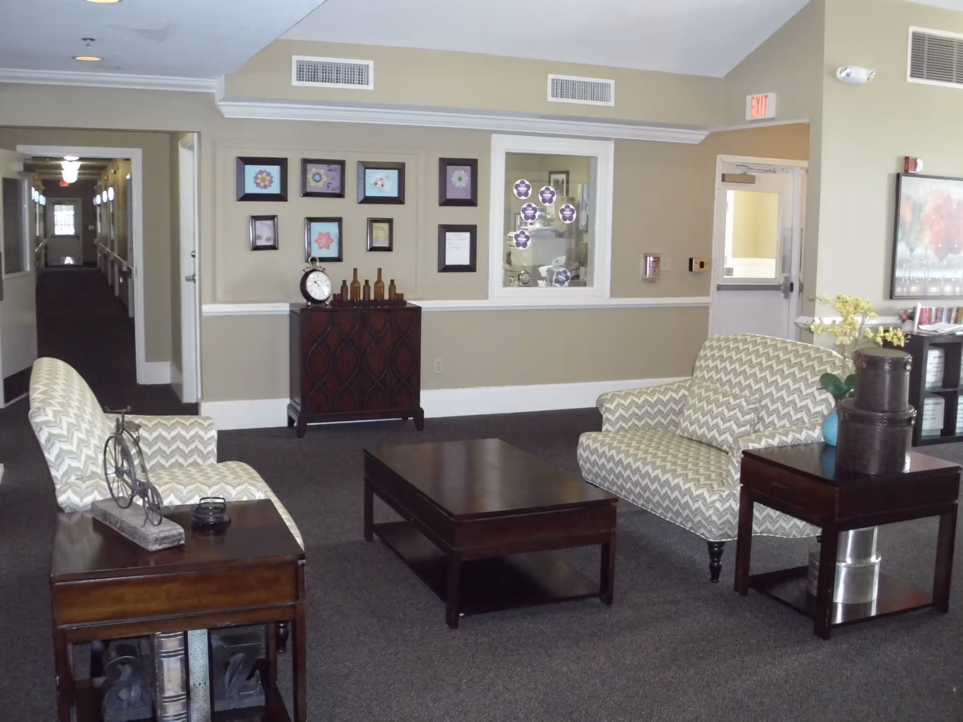 A cozy sitting area in a senior living facility with two patterned armchairs, a wooden coffee table, and two wooden side tables. One side table has decorative items including a small bicycle sculpture and books, while the other has a vase with yellow flowers and stacked containers. The walls are beige with framed artwork and a clock on a cabinet. A hallway is visible in the background, along with a door marked with an exit sign.