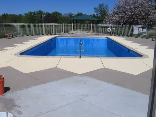 Empty outdoor swimming pool with blue interior surrounded by a beige concrete deck. There is a metal handrail in the center of the pool's edge. The pool area is fenced with a metal railing, and beyond the fence, there is a green lawn, trees, a bench, and a gazebo. The sky is clear and blue.