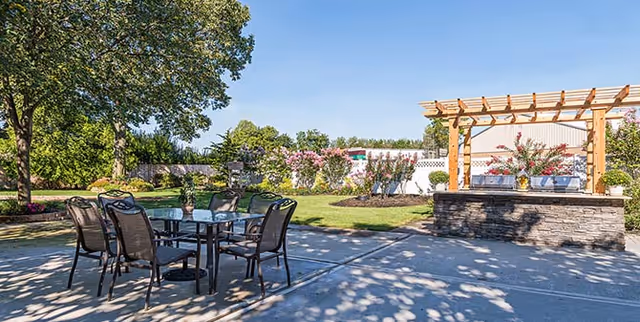 Sunny outdoor patio with a glass-top table and chairs, a pergola over a stone bar, and landscaped lawn and trees.