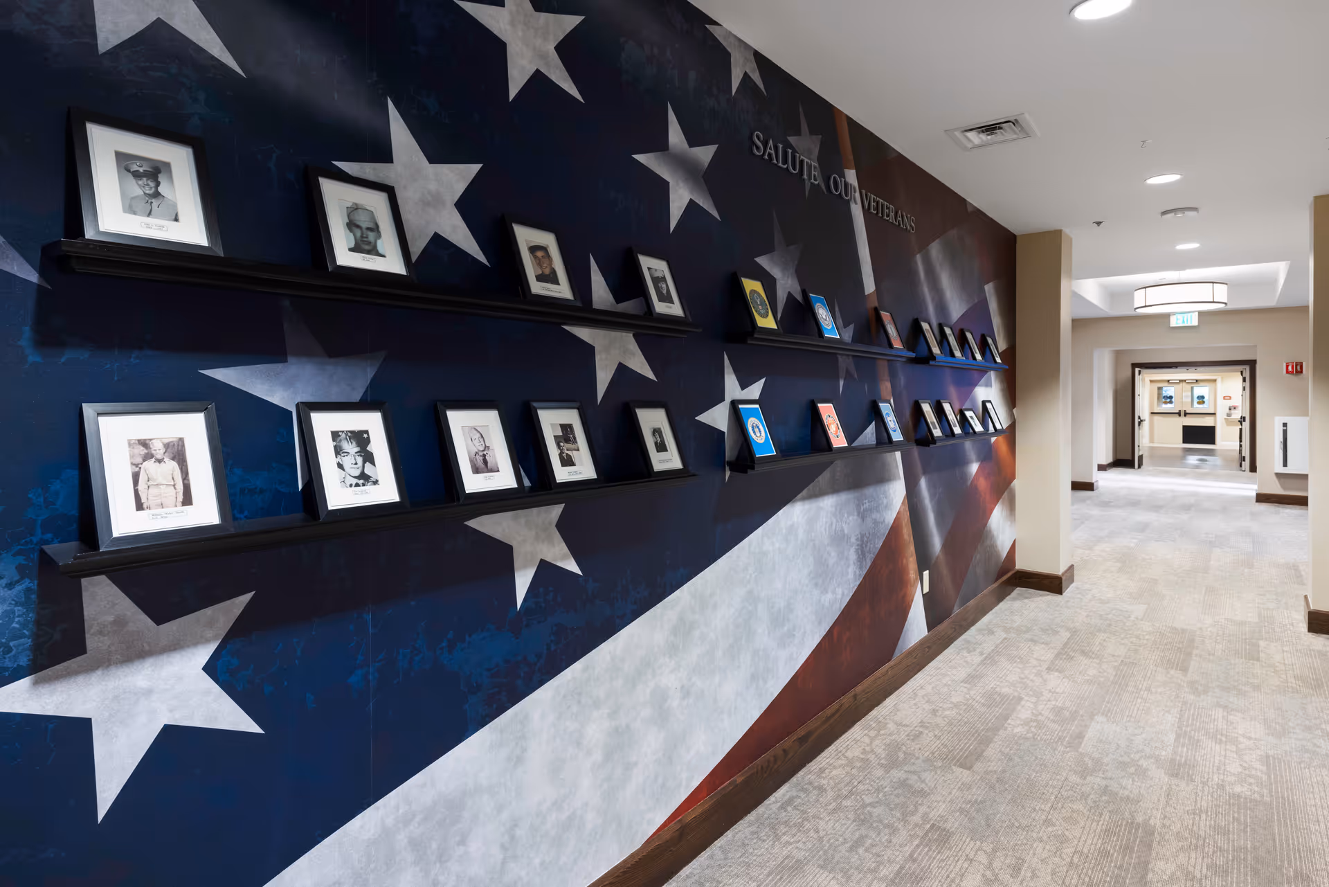 A hallway in a senior living facility with a large American flag mural on the wall. Two black shelves mounted on the wall display framed photographs and military insignia, honoring veterans. The hallway has beige walls, carpeted flooring, and ceiling lights leading to a doorway at the end.