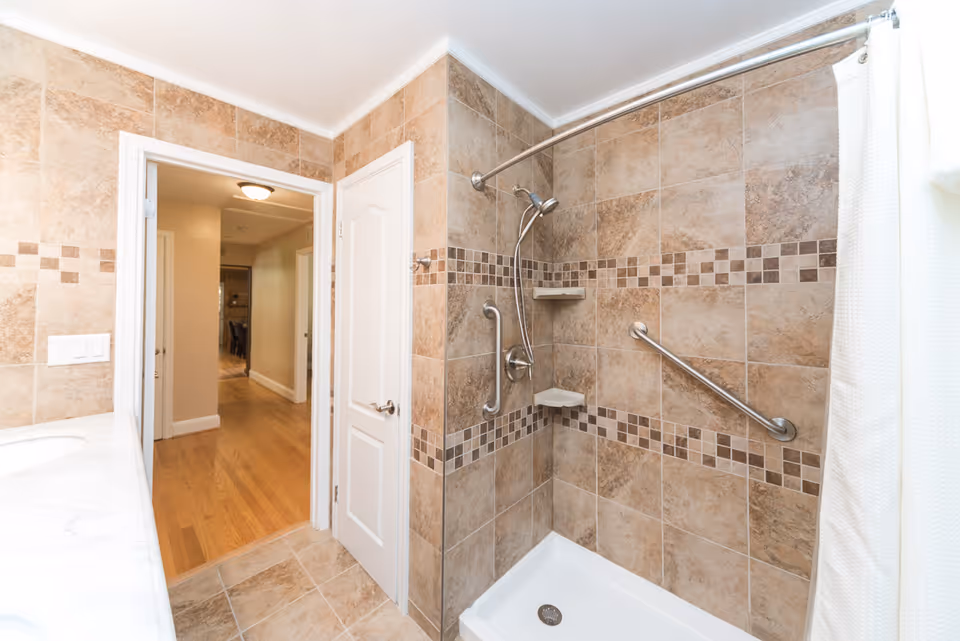 A bathroom with beige and brown tiled walls and floor, featuring a walk-in shower with a handheld showerhead, grab bars, and corner shelves. The shower has a white base and a curved shower curtain rod with a white curtain. A white door opens to a hallway with wooden flooring and beige walls.